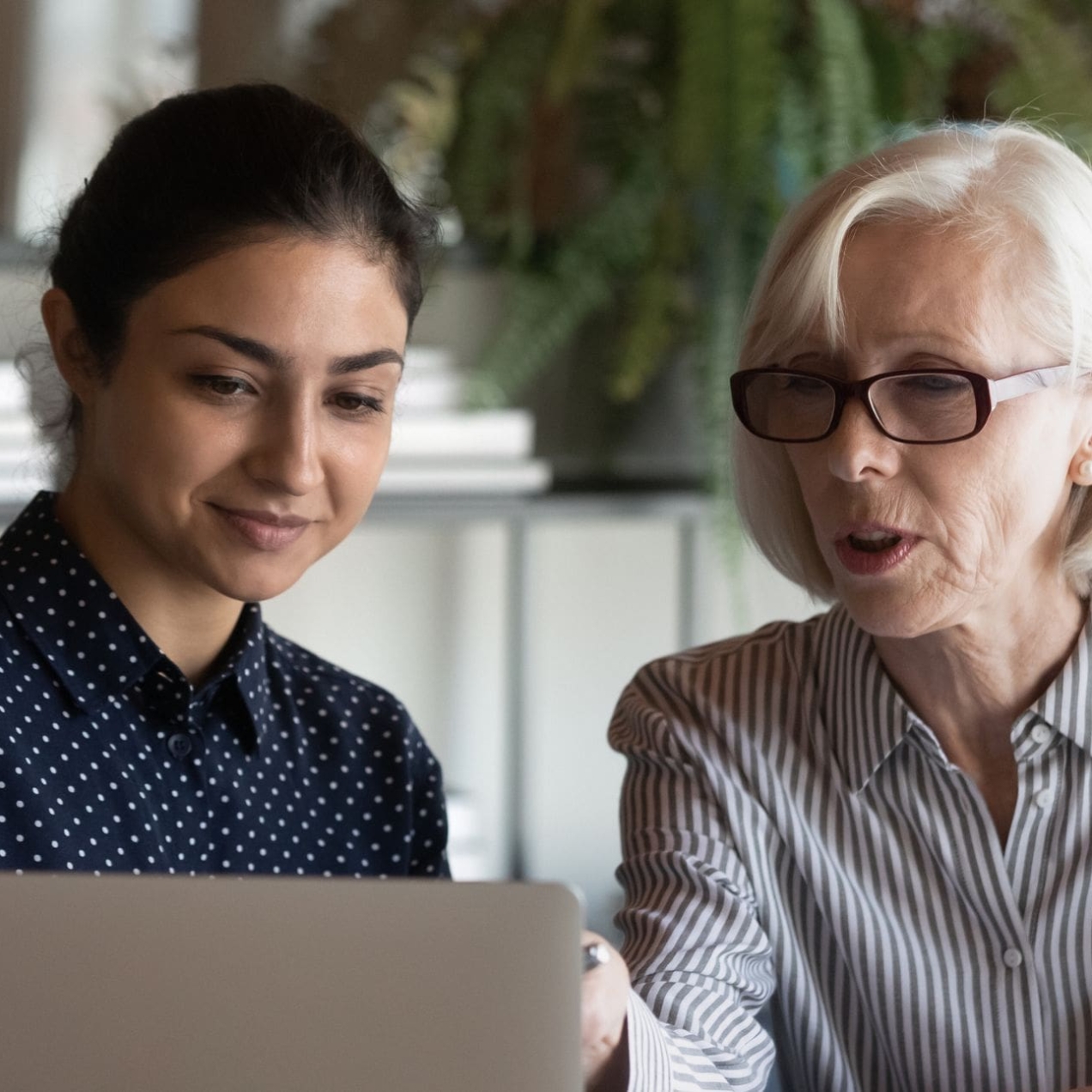 A young Asian woman with tied back dark hair and wearing a blue top, is sat looking at a laptop next to a white older woman with collar length white-bonde hair. She wears glasses and a light shirt. They are sat in an office environment in front of pl